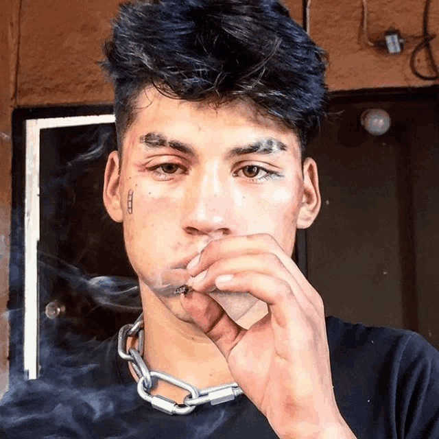 a young man wearing a chain around his neck is smoking a cigarette