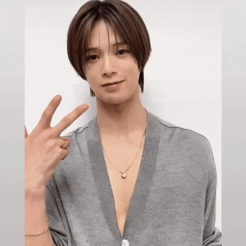 a young man wearing a grey cardigan and a gold necklace is making a peace sign