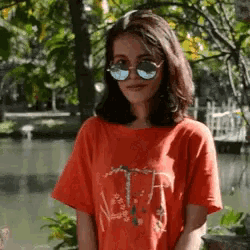 a woman wearing sunglasses and a red t-shirt stands in front of a body of water .