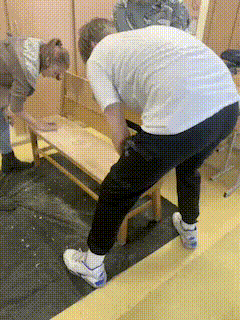 two men are working on a wooden bench and one is wearing a white shirt
