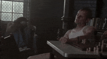 a man in a white tank top sits at a table with a chess board on it
