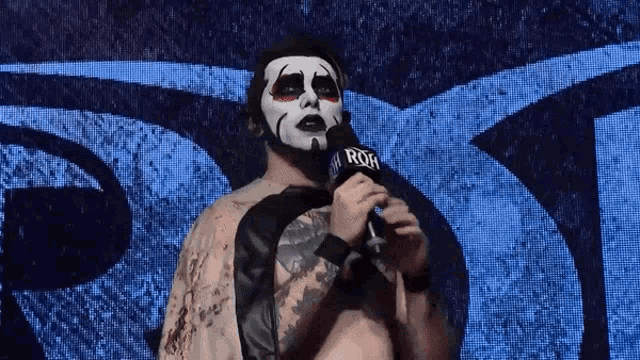 a wrestler with white face paint is holding a microphone that says roh on it