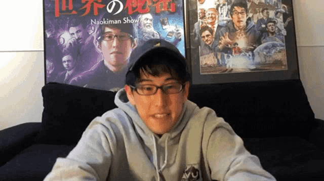 a man wearing glasses and a baseball cap is sitting on a couch in front of a naokiman shaw movie poster