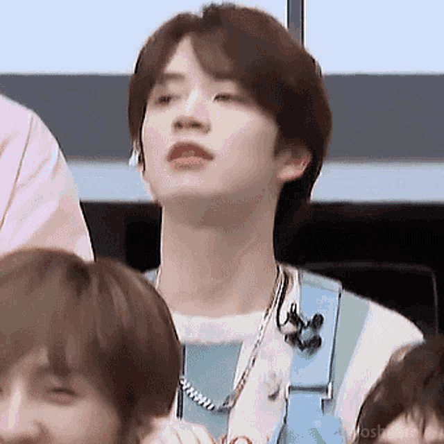 a young man wearing a necklace is sitting in a crowd of people and looking up .