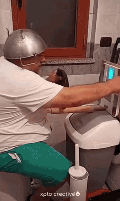a man wearing a strainer on his head is sitting on a toilet next to a trash can