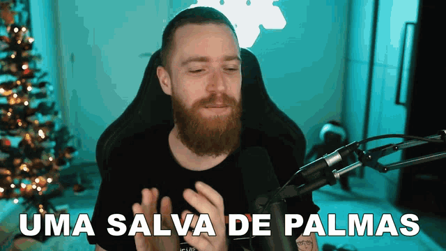 a man with a beard is sitting in front of a microphone with the words uma salva de palmas behind him