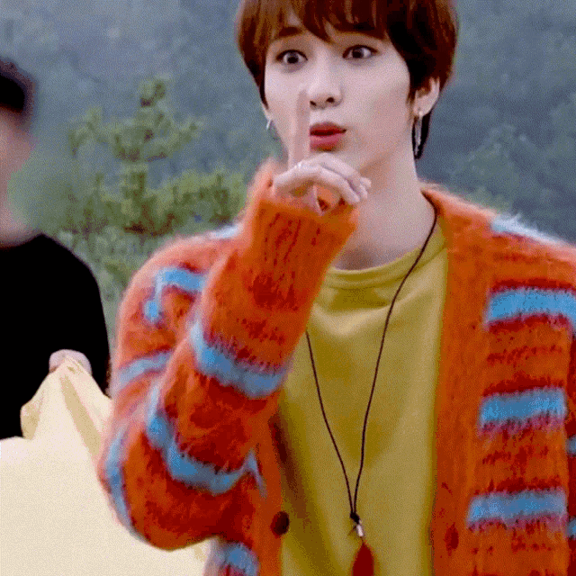 a young man wearing a yellow shirt and an orange cardigan holds his finger to his lips