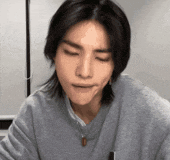 a young man with long black hair is wearing a grey sweater and a necklace .