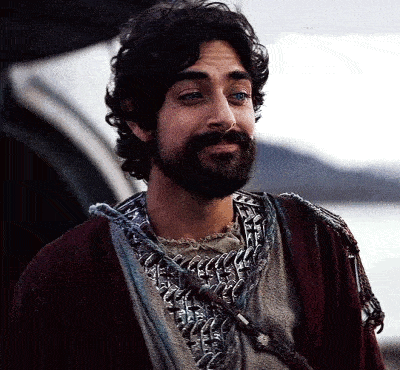 a man with a beard is standing in front of a body of water wearing a chain mail jacket .