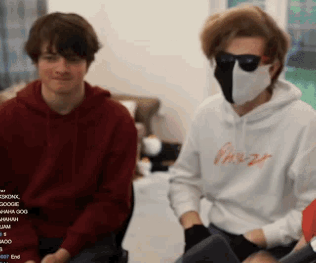 a man wearing a mask and sunglasses sits next to another man wearing a red hoodie .