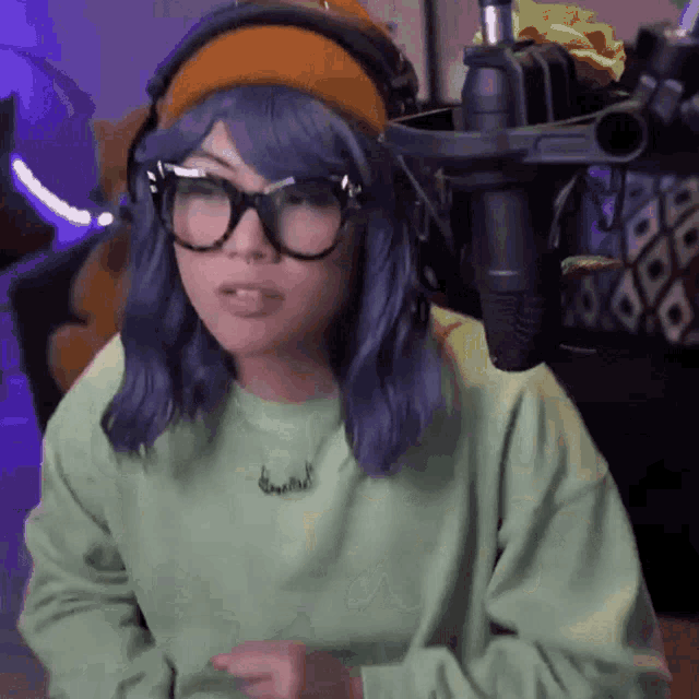 a woman with blue hair and glasses is standing in front of a microphone wearing headphones and a headband .