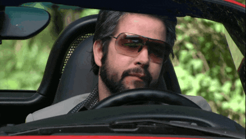 a bearded man wearing sunglasses is driving a red car