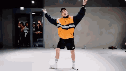 a man in an orange jacket and black shorts is dancing in a dance studio with his arms in the air .