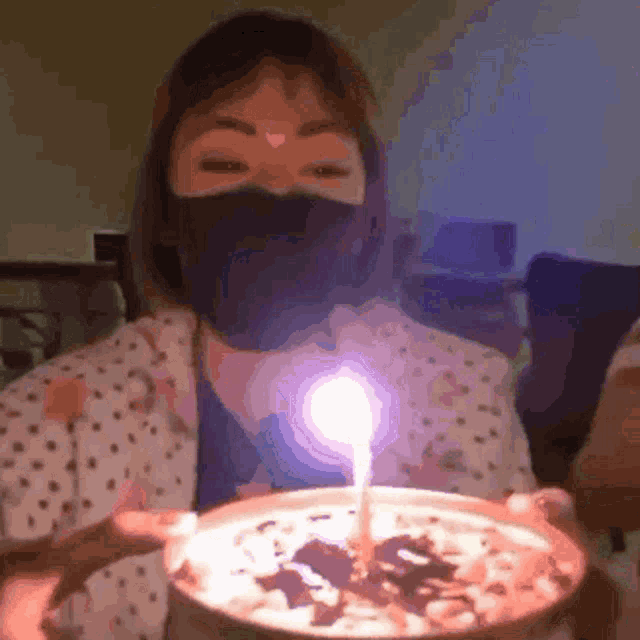a woman wearing a mask is holding a bowl of food with a lit candle in it .