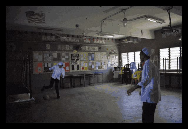 a man in a white shirt is kicking a soccer ball in a classroom with an american flag on the wall