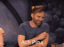 a man with a beard is sitting at a table with his arms crossed and the words ach pina behind him