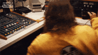 a woman in a yellow jacket sits at a desk in front of a son of a glitch mixer