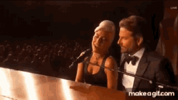 a man and a woman are singing into microphones while sitting at a piano in front of a crowd .