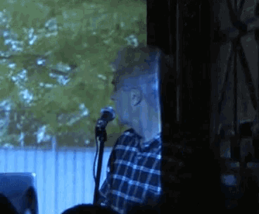 a man in a plaid shirt is singing into a microphone in front of a screen