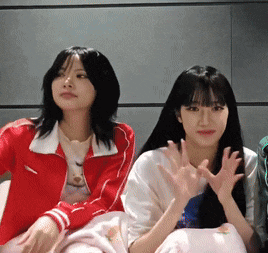 two girls are sitting next to each other on a bed and one is wearing a red jacket .