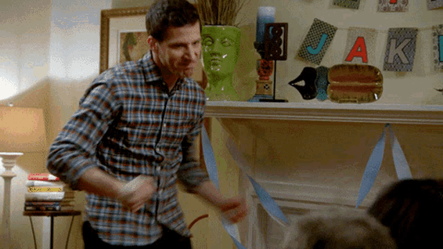 a man in a plaid shirt is dancing in front of a fireplace with the letters j and k on the wall behind him