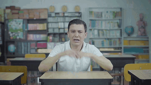 a man in a white shirt is sitting at a desk in a classroom making a funny face