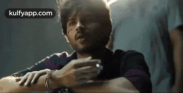 a man in a purple shirt is sitting down with his hands folded and talking to someone .