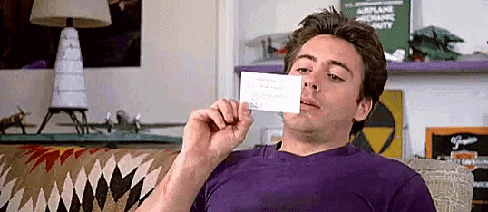 a man in a purple shirt is sitting on a couch holding a business card in front of his face .
