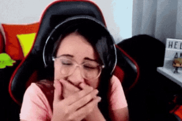a woman wearing headphones and glasses is sitting in a chair covering her mouth with her hands .