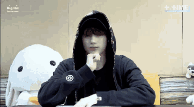 a man in a black hoodie is sitting at a table with a stuffed animal in the background