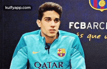 a man wearing a blue qatar jacket is sitting in front of a fc barcelona banner .