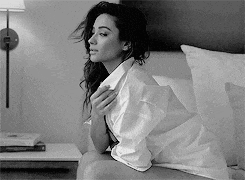 a black and white photo of a woman sitting on a bed wearing a shirt .