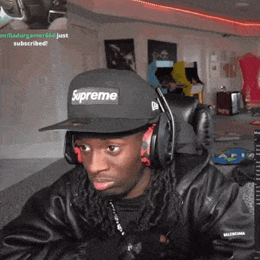 a man wearing a supreme hat and headphones sits in a chair