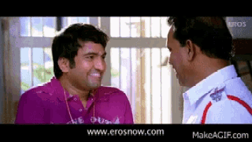 two men are talking to each other in front of a window and the website www.erosnow.com is displayed
