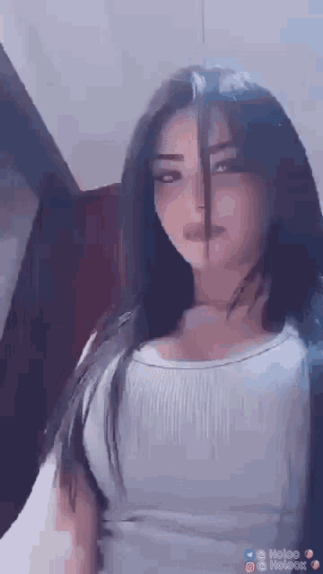 a woman with long black hair is wearing a white tank top and taking a selfie .