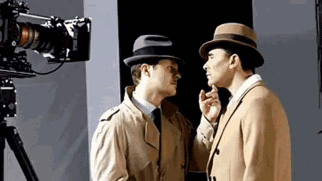 two men in suits and hats are standing next to each other in front of a camera and talking to each other .