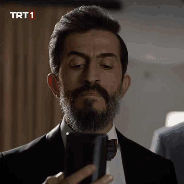 a man with a beard is looking at a cell phone with a trt1 logo in the corner
