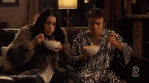 a man and a woman are sitting on a couch eating from bowls of food .