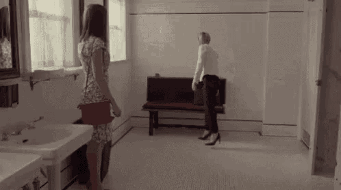 two women are standing in a bathroom next to a sink and bench .