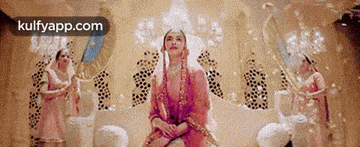 a woman in a pink dress is sitting on a throne in a room surrounded by dancing women .