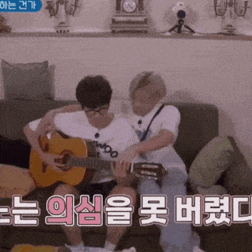 two men are sitting on a couch playing guitars and one of them has glasses on .