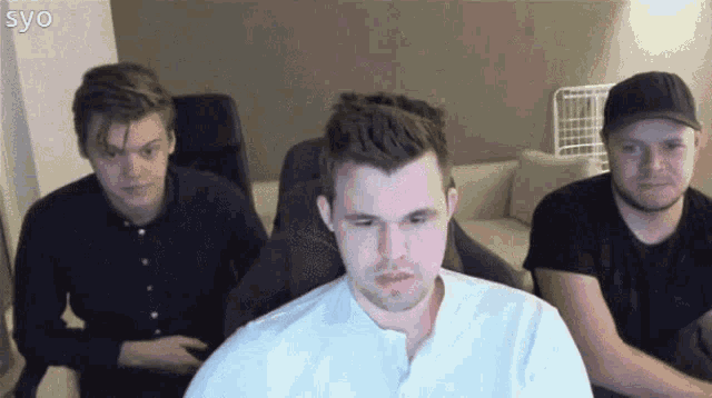 three men are sitting in chairs in front of a computer screen and one of them is wearing a white shirt .