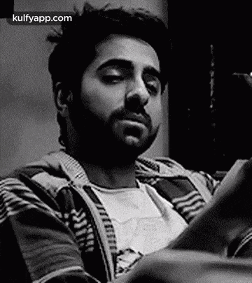 a man with a beard is sitting on a couch looking at a cell phone .