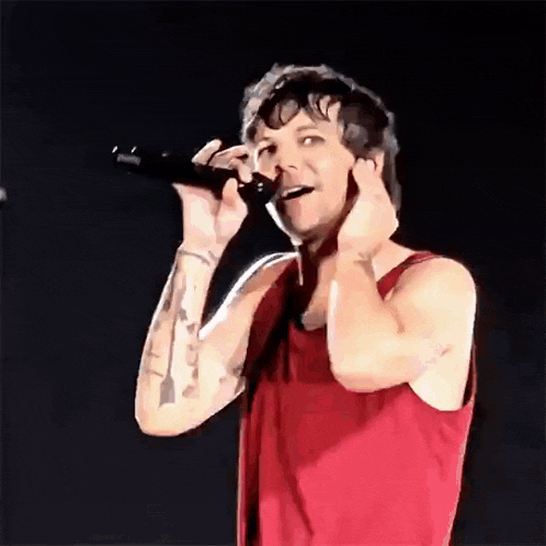 a man in a red tank top is singing into a microphone while covering his ears .