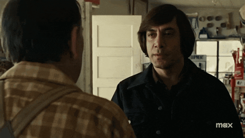 a man in a plaid shirt is talking to another man in a room with a max logo on the bottom