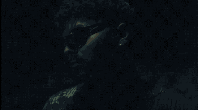 a man wearing sunglasses looks at the camera in a dark room