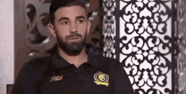 a man with a beard wearing a black shirt is sitting in front of a wall .
