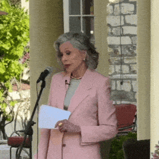 a woman in a pink suit stands in front of a microphone holding a piece of paper