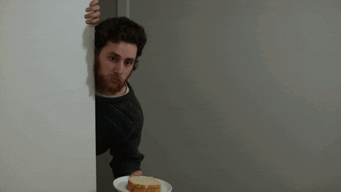a man peeking out from behind a wall holding a plate with a piece of bread on it