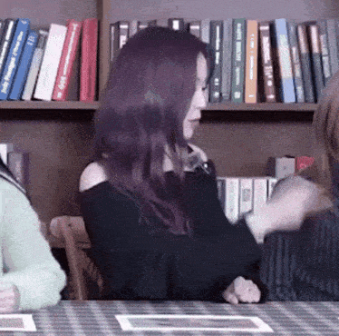 a woman with purple hair is sitting at a table in front of a bookshelf filled with books